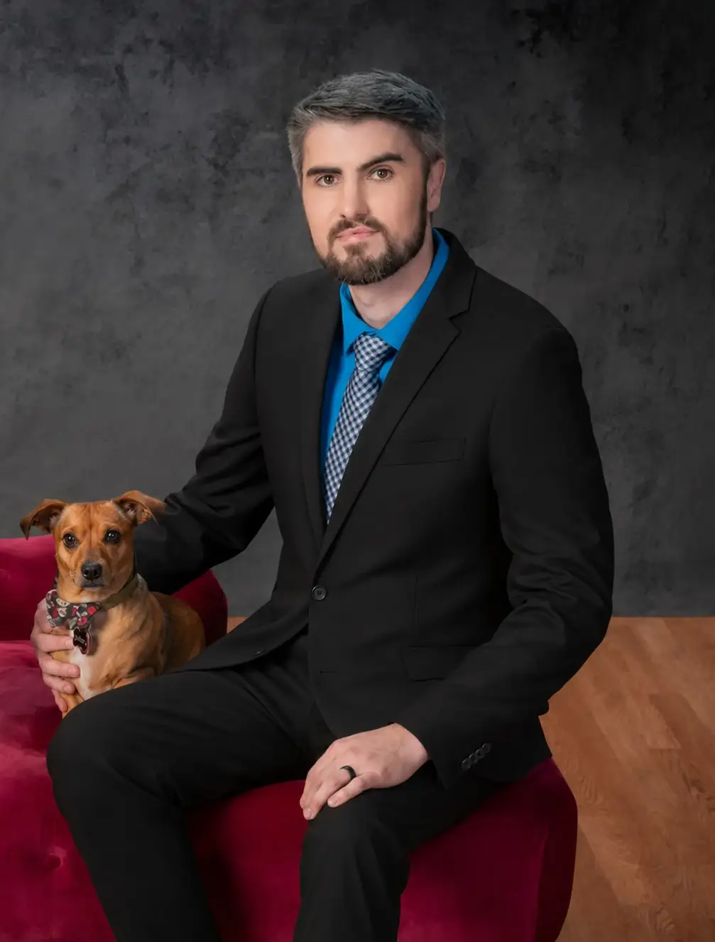 Dieter Grosswiler in a dark suit and blue shirt, seated on a deep red velvet couch with his dog Dash